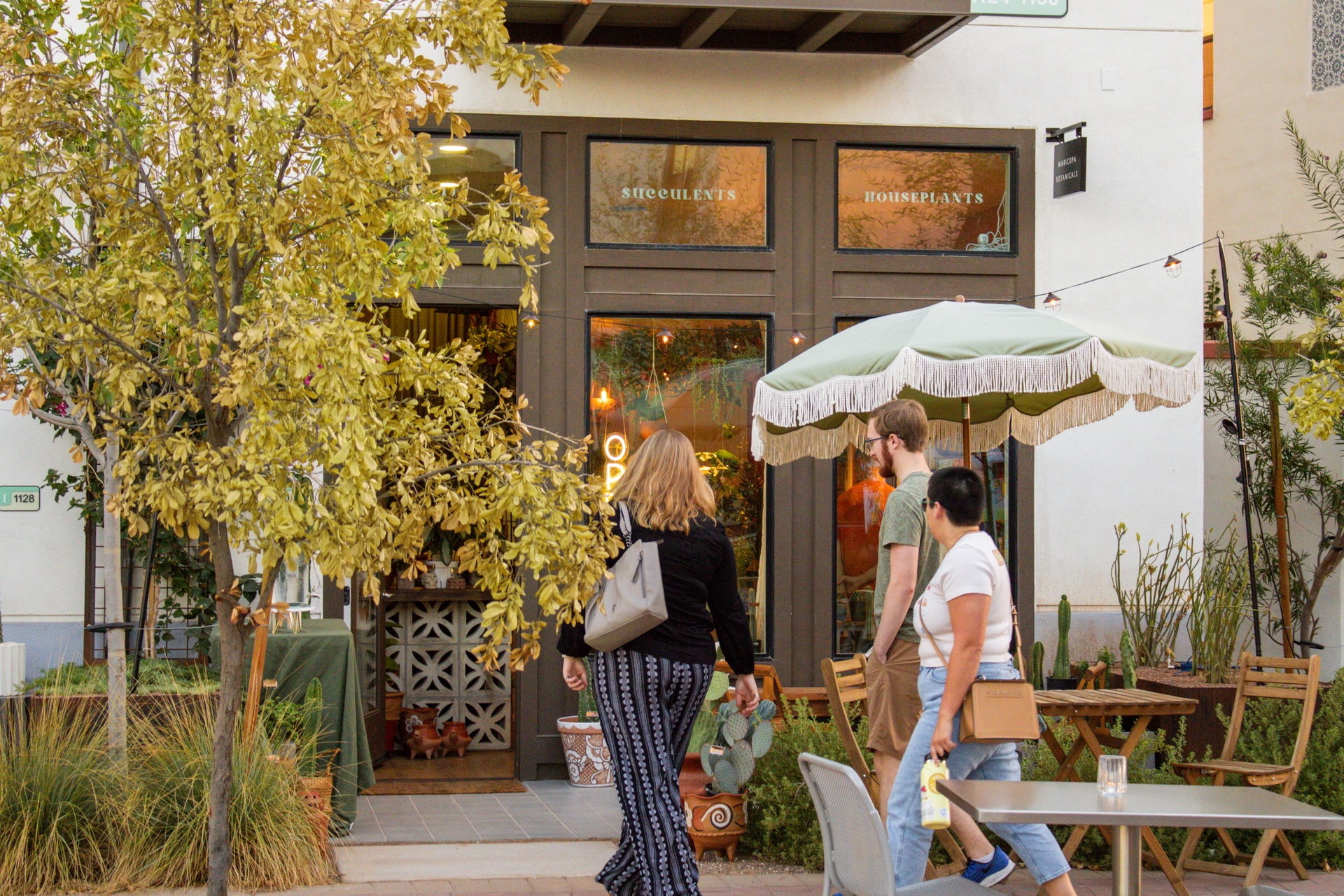 Three people about to walk into a shop with a tree whose leaves are starting to turn color